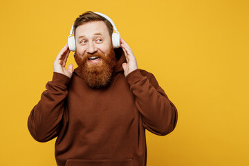 Young cheerful fun happy redhead caucasian man wearing brown hoody casual clothes headphones listen to music look aside on area isolated on plain yellow background studio portrait. Lifestyle concept.