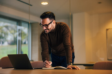 Male employer looking at the laptop, taking notes, and leaning on the desk at the office.