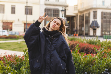 Wall Mural - Cool blonde woman in dark blue bomber shows peace v sign, closed eyes, blows kiss and posing outside. Happy woman walk and posing outdoor in the city.