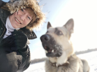 An adult girl or a middle aged woman in jacket with a fluffy fur hood with a shepherd dog in nature in winter on a cold sunny day