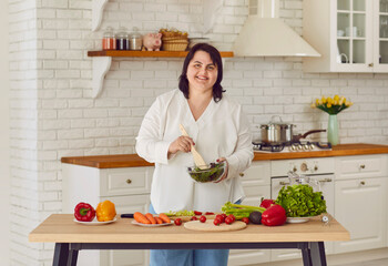 Smiling overweight woman preparing fresh vegetable salad in kitchen. Positive plus size young woman cooking healthy food for dinner at home. Healthy eating, organic vegetarian food