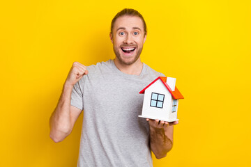 Poster - Photo of positive lucky man wear grey t-shirt holding small house rising fist isolated yellow color background