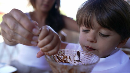 Cute little boy eating chocolate ice cream with spoon. Closeup face of male kid eats sweet dessert reward with napkin around neck