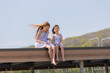 © Tina kids photo - two happy little girls are sitting on a wooden pier on a city lake in summer. Blue sky. Earth Day. Children's Day.Space for text