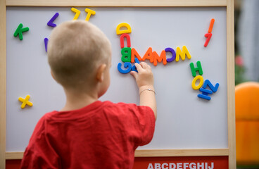 Back view of a blond toddler boy playing with colorful letters on a magnetic board