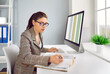 © Studio Romantic - Business accountant working in office. Serious busy woman in suit and glasses sitting in front of computer screen, checking book of financial records and using spreadsheet files on PC