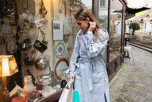 Young Woman In Trench Coat Standing With Shopping Bags And Looking At Showcase Of Antique Shop In Vienna.