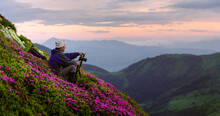Photographer In The Spring Forest During Sunset. Taking Photo Of Rhododendron Flowers Covered Mountains Meadow. Purple Sunrise Light Glowing On A Foreground. Landscape Photography