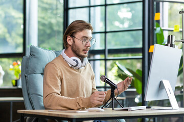 A young male blogger is sitting in a home office at a desk with a computer in front of a microphone and wearing headphones. Records podcast, blog, communicates online via video call, webinar.