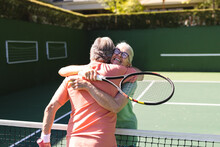 Happy Caucasian Senior Friends With Tennis Rackets Embracing At Tennis Court On Sunny Day
