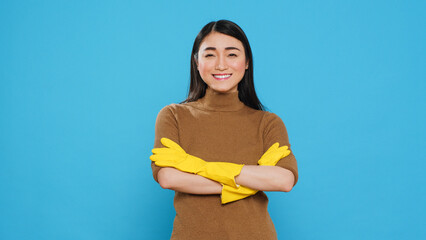 Wall Mural - Smiling professional maid wearing protective rubber gloves standing with arm crossed in studio over blue background. Cheerful asian housekepper took great care in customer housecleaning duties