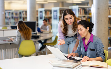 Wall Mural - Two woman students, who are preparing a report on a laptop in the university library, discuss studying issues