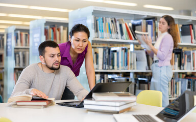 Wall Mural - Interested young bearded man working with his asian female colleague on joint research in public library, looking for information in books and Internet ..