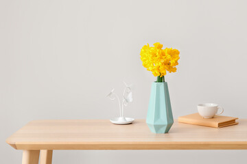 Vase with narcissus flowers, book and cup of tea on table near light wall