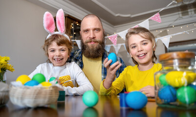 Easter Family traditions. Father and two caucasian happy children with bunny ears dye and decorate eggs with paints for holidays while sitting together at home table. Looking at camera