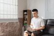 © Anciens - Asian man is using laptop at home, sitting on the couch.