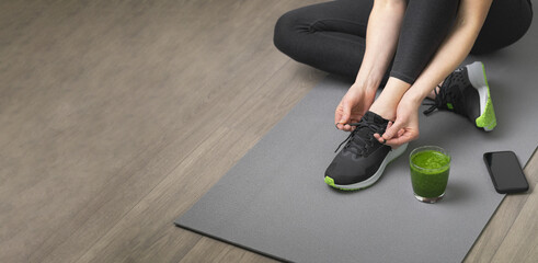 Women's hands tying sport shoes on a gray workout mat. With smoothie for detox in background. Healthy living, dieting lifestyle. Banner