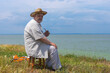 Outdoor portrait of chubby Ukrainian senior man looking into the distance while sitting on wicker small stool on Dipro riverside at spring season