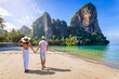 A happy couple on holidays walks down the empty Railay Beach at Krabi, Thailand, with emerald sea and lush rain forest