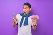 © Bangun Stock Photo - Portrait of shocked young Asian man using mobile phone and holding money banknotes isolated over purple background