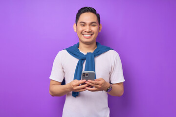 Wall Mural - Cheerful young Asian man holding smartphone and smiling at camera isolated over purple background