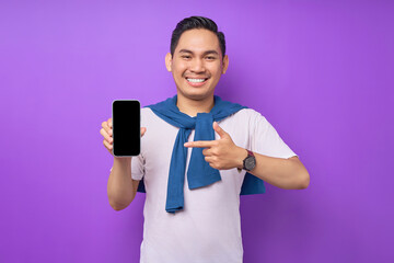 Wall Mural - Cheerful young Asian man pointing index finger at empty smartphone screen isolated on purple background. Advertising new mobile app, mockup concept