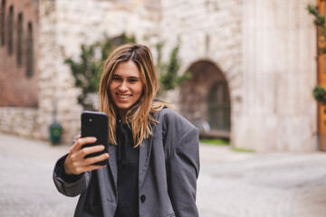 Wall Mural - Happy young woman photographing herself using her mobile phone. Caucasian female talking selfie with her smart phone walking outdoor on the street. Girl posing in grey suit, t-shirt.