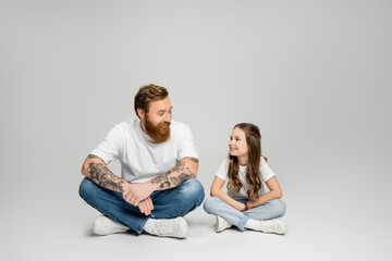 Poster - Smiling girl talking to tattooed father while sitting on grey background.