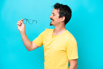 Wall Mural - Young caucasian man holding glasses isolated on blue background with happy expression