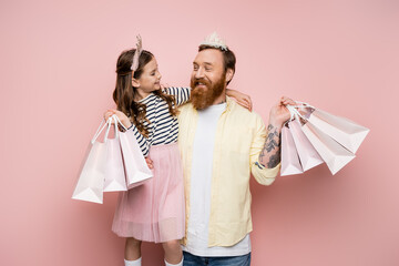 Poster - Cheerful girl holding shopping bags and hugging father with crown headband on pink background.