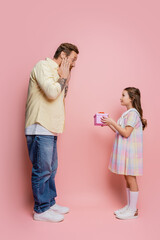 Poster - Side view of shocked man looking at preteen daughter with gift on pink background.