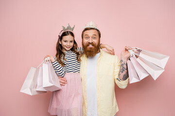 Wall Mural - Cheerful man and daughter with crown headbands holding shopping bags on pink background.