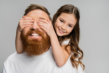 Wall Mural - Cheerful preteen girl covering eyes of happy father with beard isolated on grey.