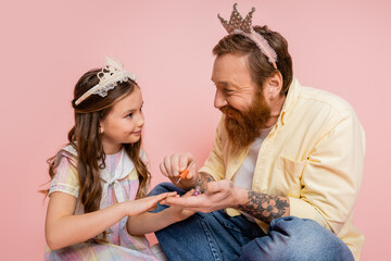 Wall Mural - Cheerful man with crown on head holding nail polish near preteen daughter on pink background.