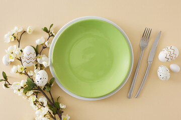 Wall Mural - Easter decor idea. Flat lay photo of empty green plate white golden easter eggs cutlery fork knife spring blossom branch on isolated beige background