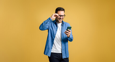 happy young adult man wearing blue denim shirt adjusting eyeglasses and text messaging over mobile p