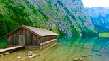 Fototapeta Góry - Konigssee, Germany - June 16, 2018: side view of lakeside cabin or traditional wooden boat house in Obersee on the left with a peaceful and clear lake on a sunny summer day