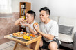 © Anciens - Asian man is eating pizza with friend in the living room.