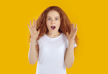 Shocked amazed young curly redhead girl with long hair in white t-shirt with raised hands and opened mouth on yellow background. Sincere human emotions concept, banner for advertisement and marketing.