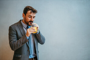 Wall Mural - A handsome businessman standing against the gray background and drinking fresh orange juice. Copy space