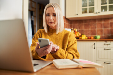 Wall Mural - Mature woman at home calculating home finances with smartphone and laptop in kitchen