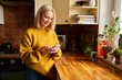 © baranq - Smiling mature woman using phone texting message while standing in a kitchen