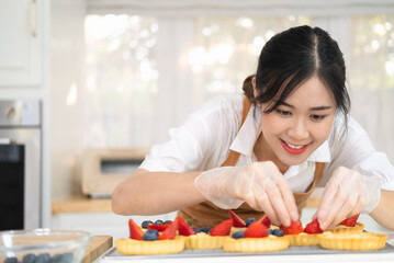 Wall Mural - Happy young housewife hands in cooking gloves decorating freshly baked tart with blueberries strawberry fresh fruit.Woman chef baker wear apron making fruit tart. homemade bakery at home.