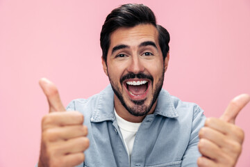 Fashion portrait of a brunette man with a beard showing a thumbs up sign on a pink background in a white T-shirt with a smile and joyful emotion on his face, copy space