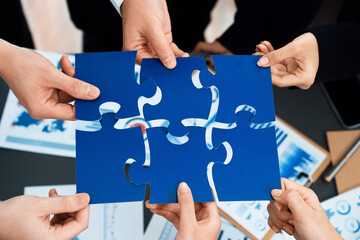 Closeup top view business team of office worker putting jigsaw puzzle together over table filled with financial report paper in workplace with manager to promote harmony concept in meeting room.