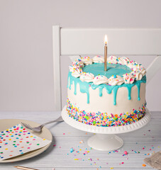 Wall Mural - Birthday party with vanilla buttercream Cake, lit gold candle, blue drip and sprinkles on a light grey white background