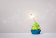 Green cupcake with sprinkles and lit sparkler on a white grey background.