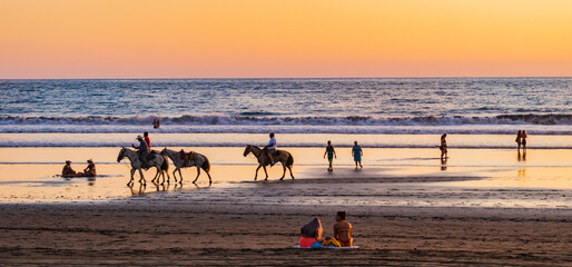 people enjoying the sunset on the beach in Jaco, Costa Rica while a cowboy leads two riders on horses 
