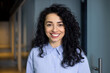© Liubomir - Close-up portrait of mature adult business woman, boss smiling and looking at camera, pleased hispanic woman with curly hair in blue shirt inside office at workplace in hall corridor.