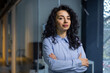 © Liubomir - Portrait of serious female boss inside business company office, businesswoman crossed arms looking concentrated at camera, wearing shirt, satisfied and successful hispanic woman with curly hair.